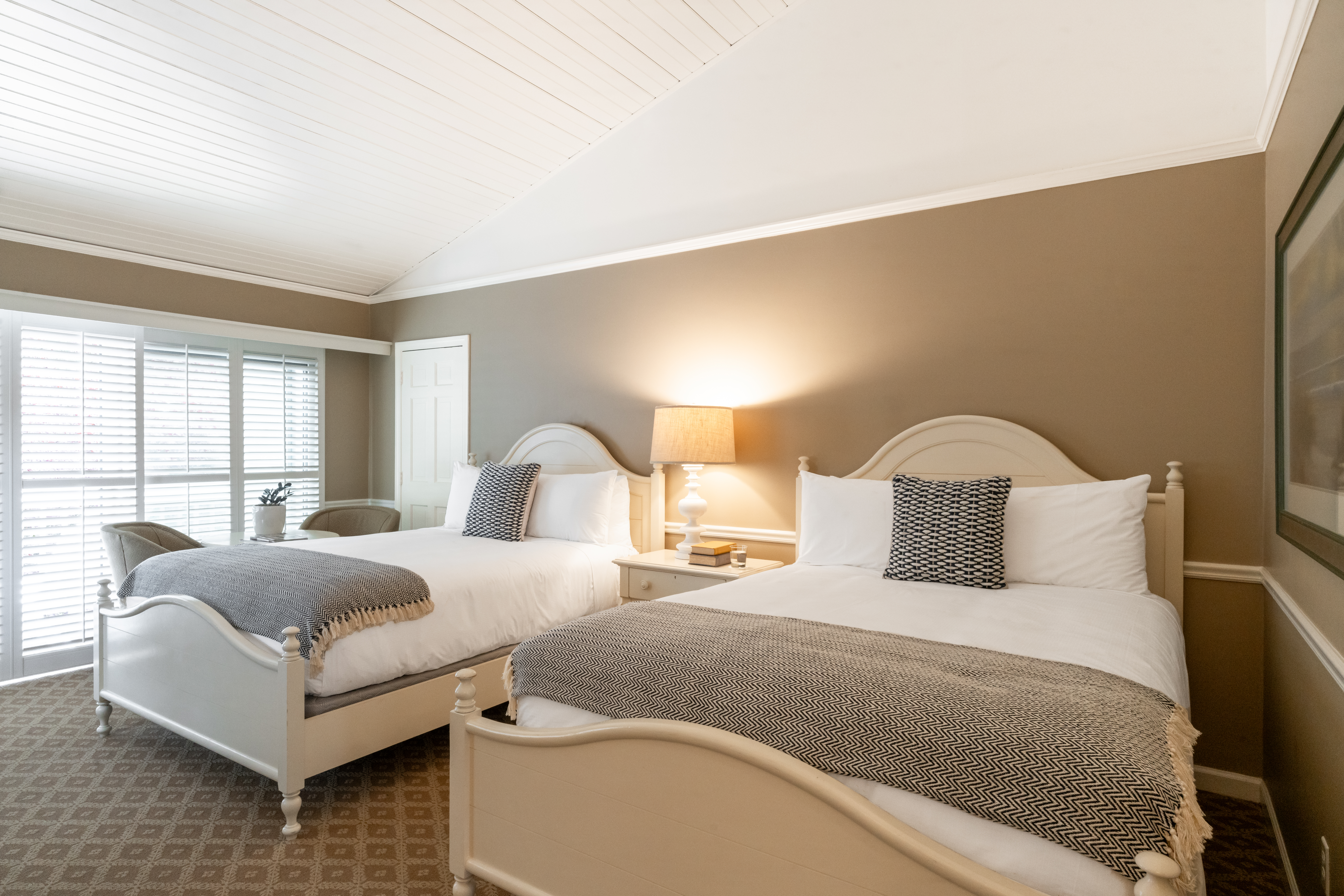 A cozy bedroom with two double beds, each with white linens and grey throws, sharing a nightstand with a lamp. The room has light brown walls, a sloped white ceiling, and a large window with blinds.