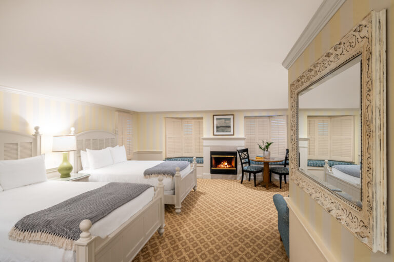 Cottage Style hotel room with two double beds, fireplace, large mirror, table with chairs, and windows with shutters. The room is decorated in light colors with patterned carpeting.