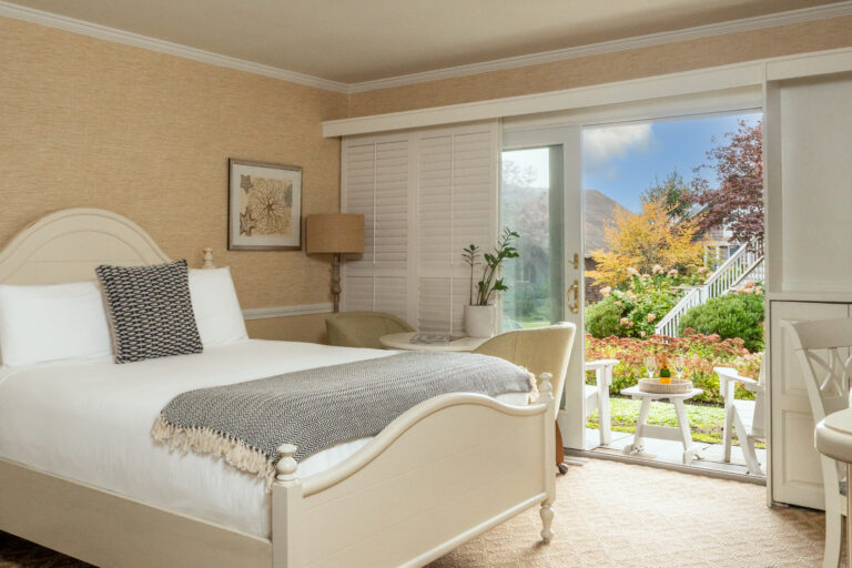 A cozy bedroom with a white bed frame, a gray blanket, an armchair, and a small table by glass doors that open to a garden with colorful foliage and a staircase.