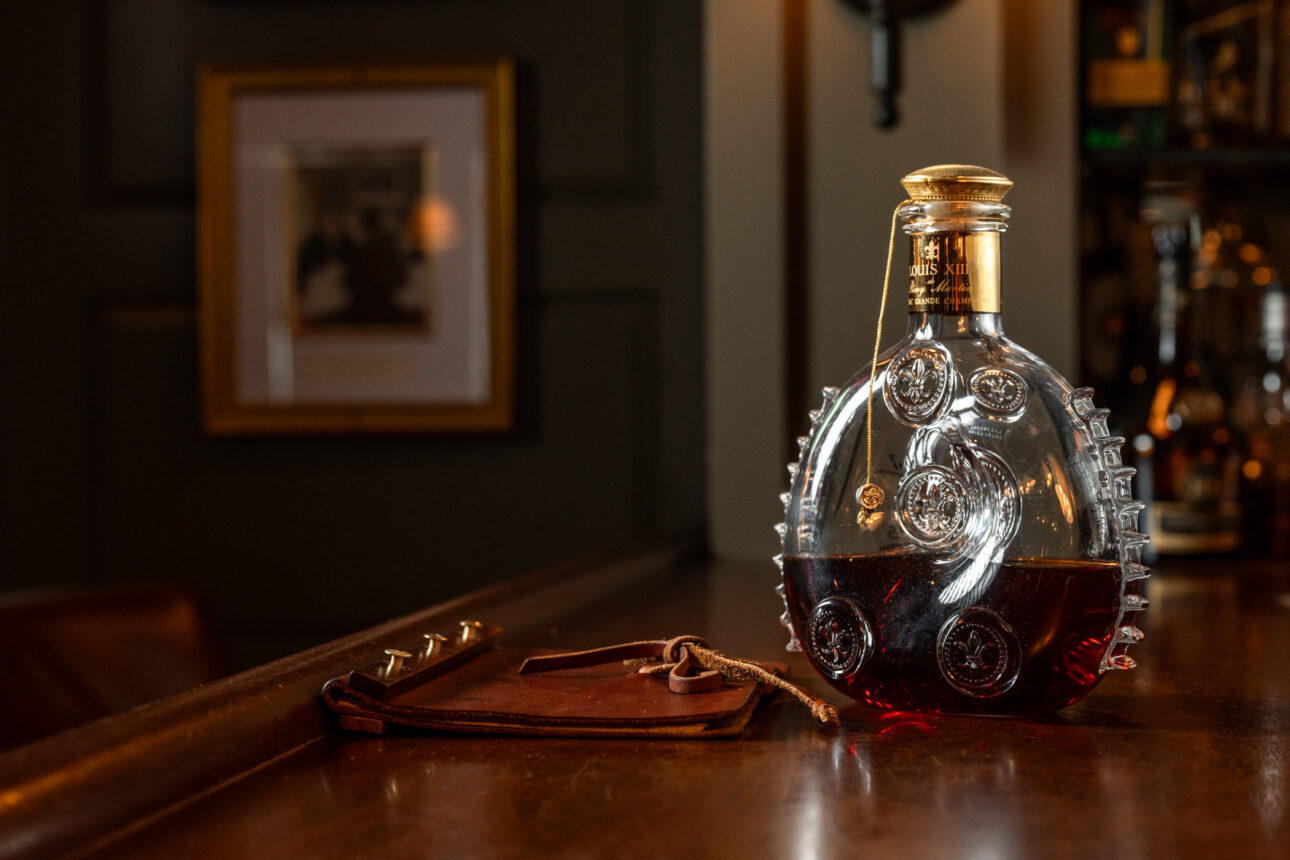 A round, ornate bottle containing brown liquid sits on a wooden bar beside a small leather pouch. The background is dimly lit with a framed picture on a dark wall.