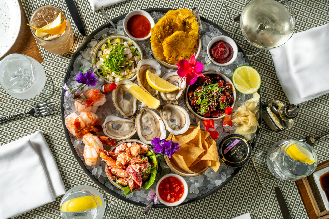 A seafood platter featuring oysters, shrimp, ceviche, and lobster, accompanied by sauces, chips, lemon wedges, and garnished with flowers, is served on a table with various drinks and napkins.
