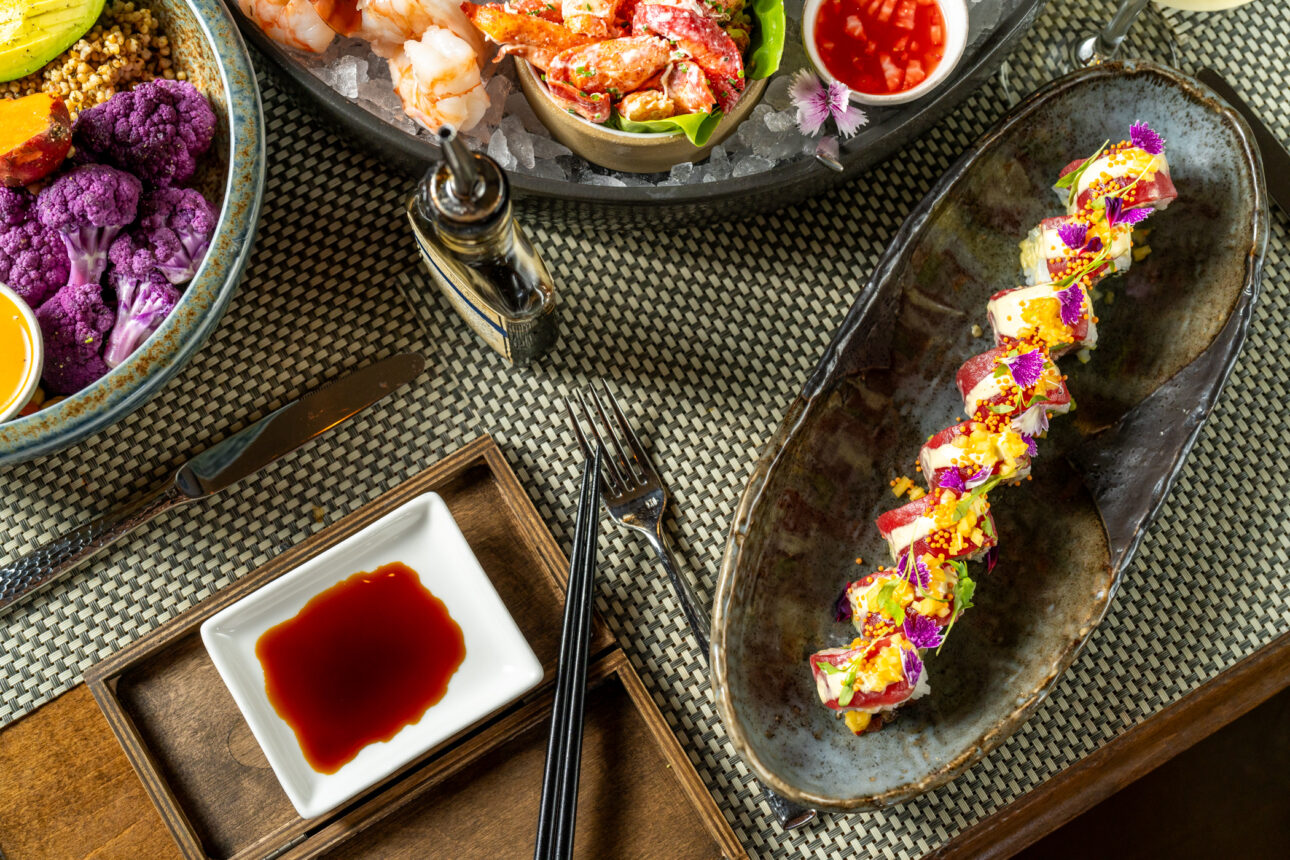A top-down view of a table with assorted dishes including sushi rolls, shrimp cocktail, and a bowl of mixed vegetables, accompanied by soy sauce and cutlery.