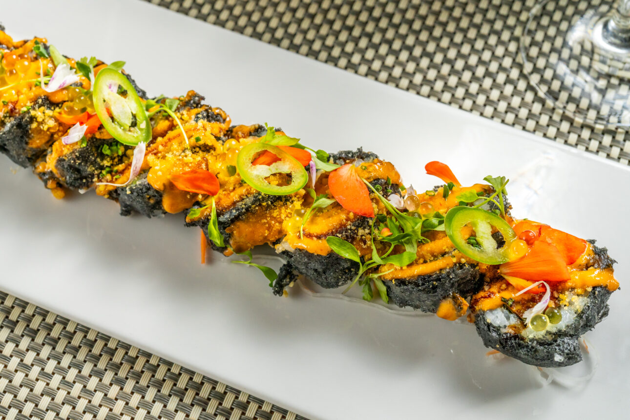 A plate of sushi rolls topped with various garnishes including sliced jalapeños, microgreens, and an orange sauce, arranged on a white rectangular dish.