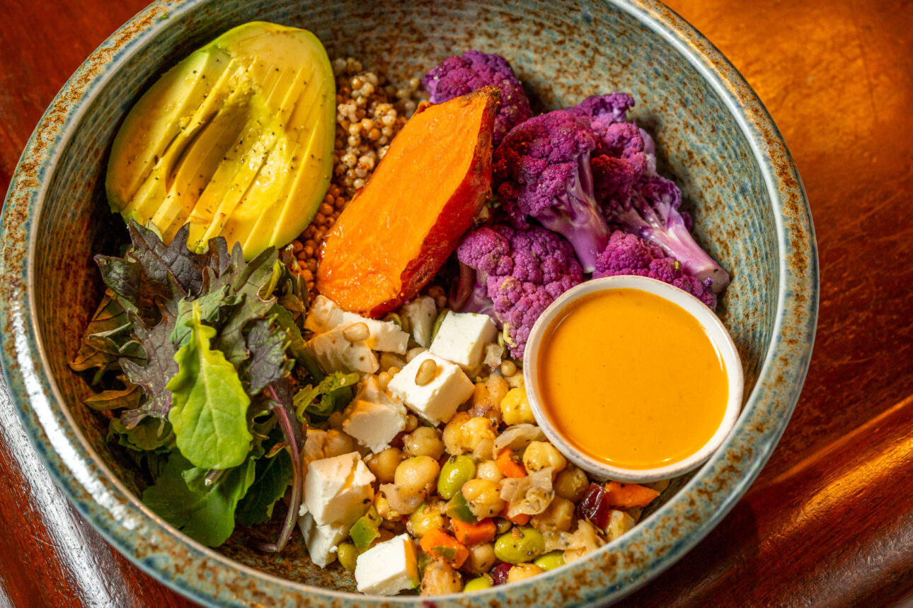 A bowl of quinoa, chickpeas, purple cauliflower, sweet potato, avocado, mixed greens, feta cheese, and a small cup of orange dressing.