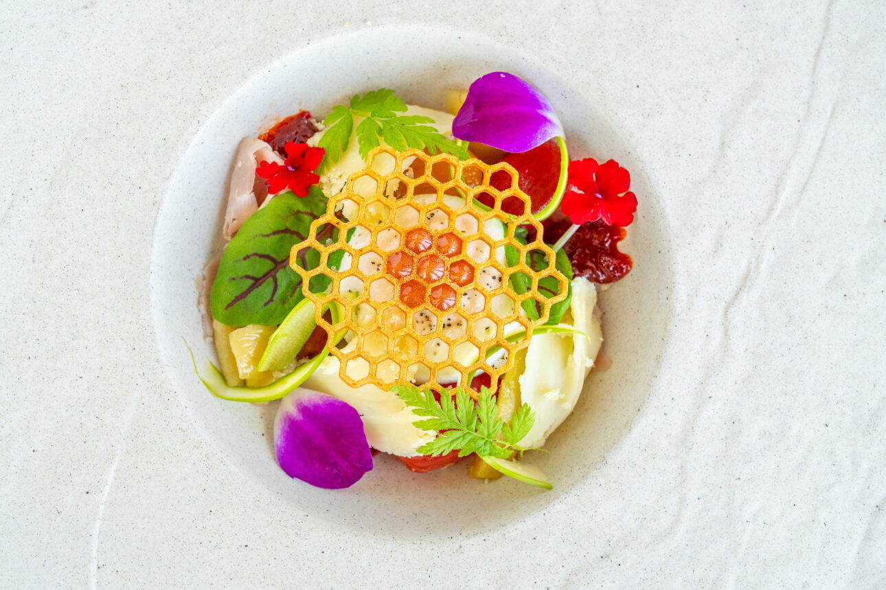 A decorative food dish features a honeycomb-shaped garnish, with assorted colorful edible flowers, greens, and other ingredients arranged artfully in a white bowl.