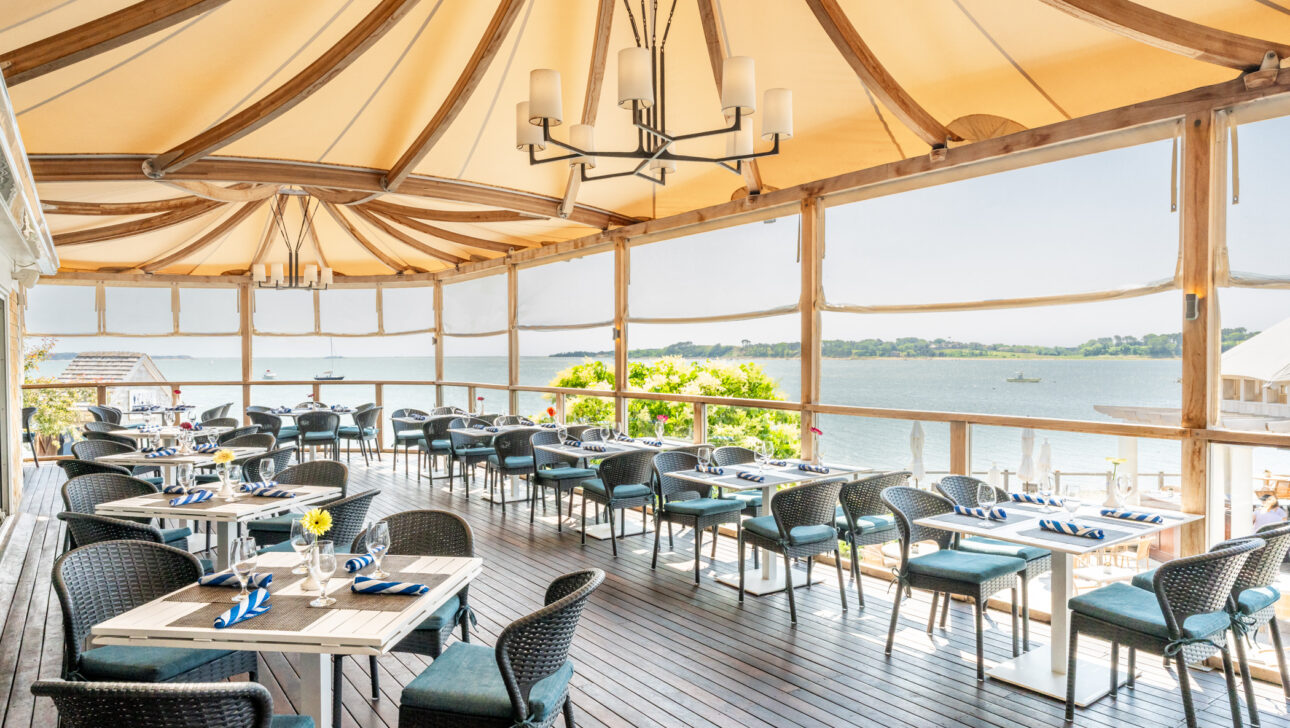 Open-air dining area with tables and chairs arranged on a wooden deck, shaded by a fabric canopy, overlooking a scenic waterfront view on a sunny day.