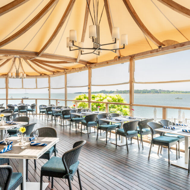 Open-air dining area with tables and chairs arranged on a wooden deck, shaded by a fabric canopy, overlooking a scenic waterfront view on a sunny day.