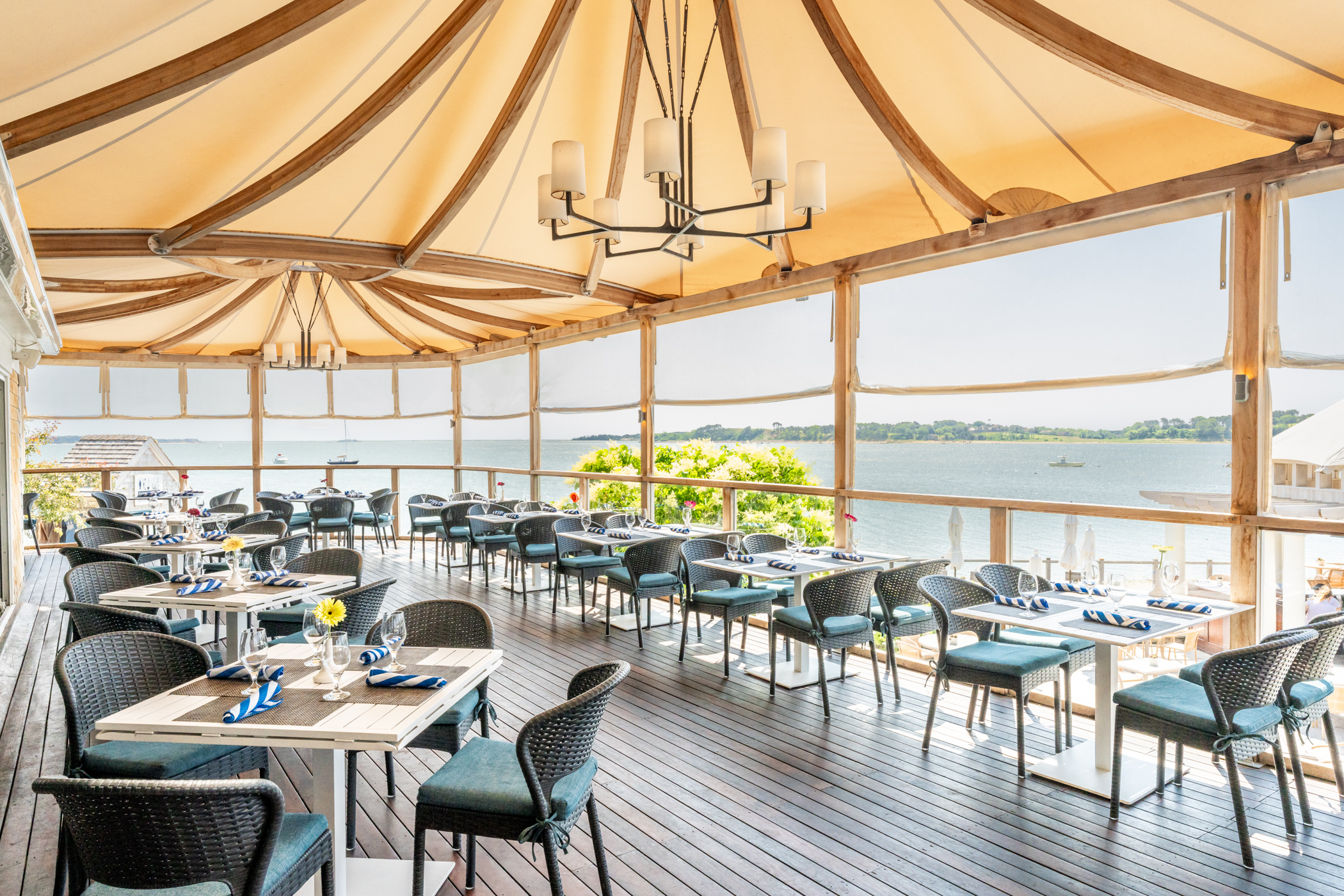 Open-air dining area with tables and chairs arranged on a wooden deck, shaded by a fabric canopy, overlooking a scenic waterfront view on a sunny day.