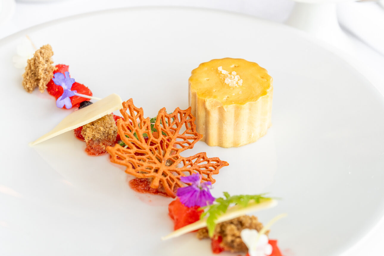 A gourmet dessert on a white plate featuring a round pastry with a golden top, accompanied by vibrant garnishes including edible flowers, fruit pieces and intricate sugar decorations.