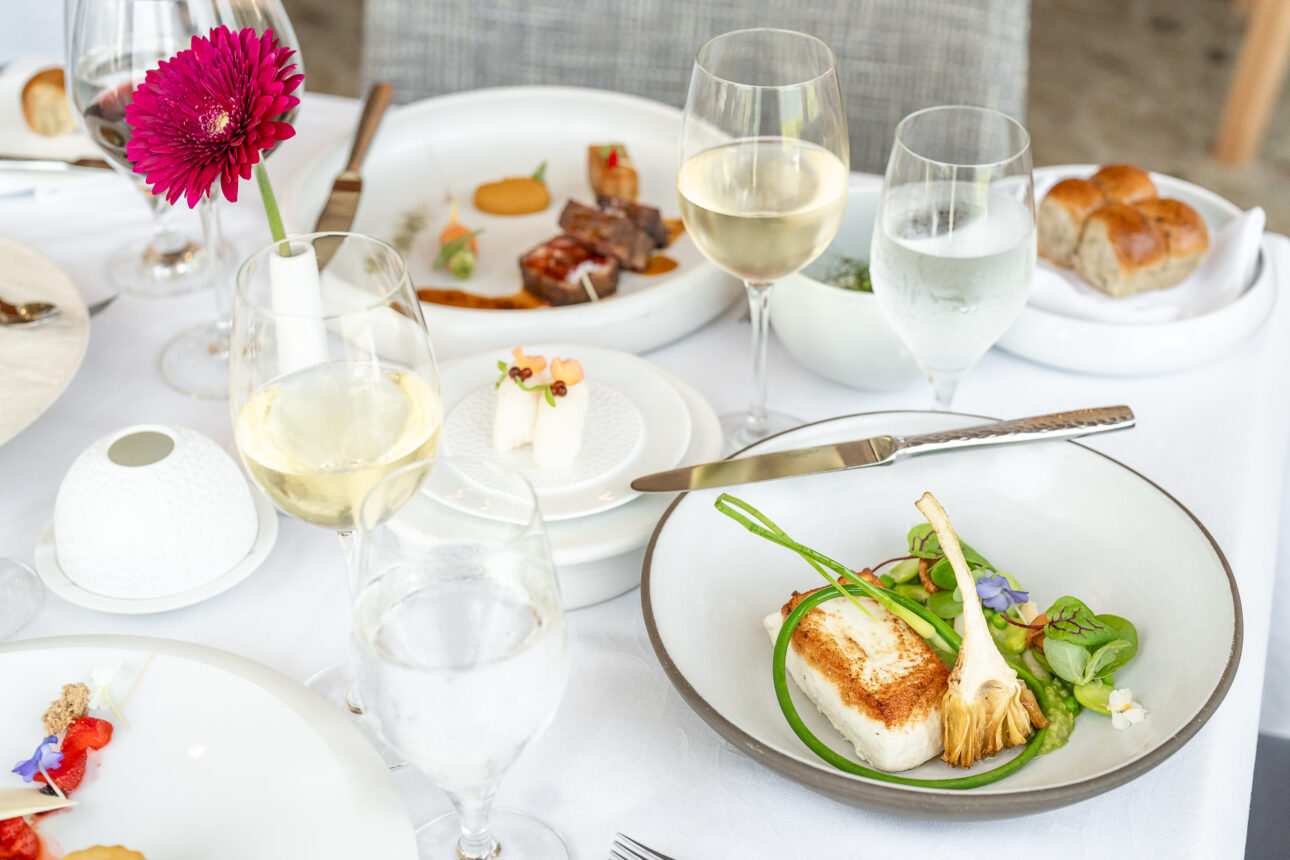 A dining table set with a variety of elegantly plated dishes, including a main course with greens and cutlery, glasses of white wine and water, and a vase with a pink flower.