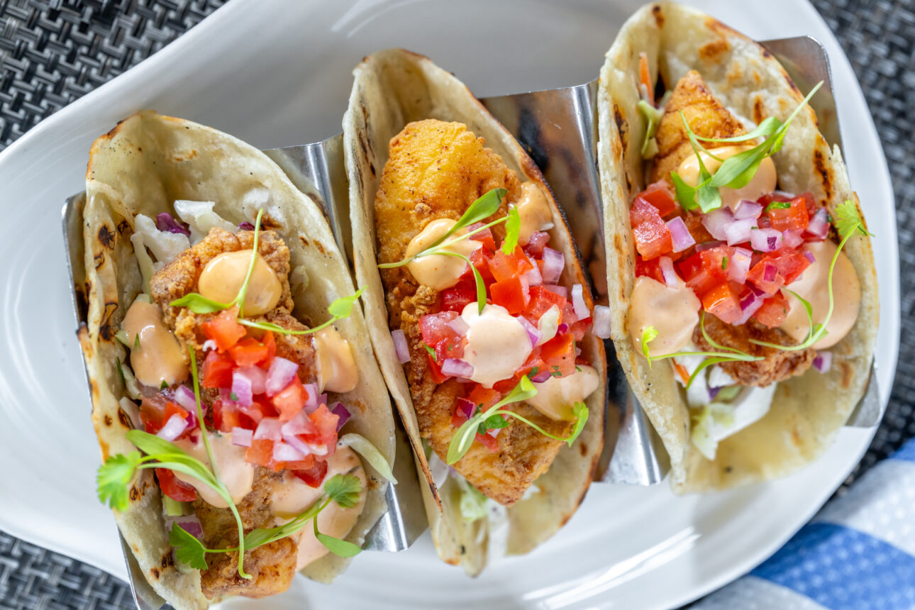Three fish tacos topped with diced tomatoes, onions, microgreens, and a drizzle of creamy sauce are placed on a white ceramic plate.
