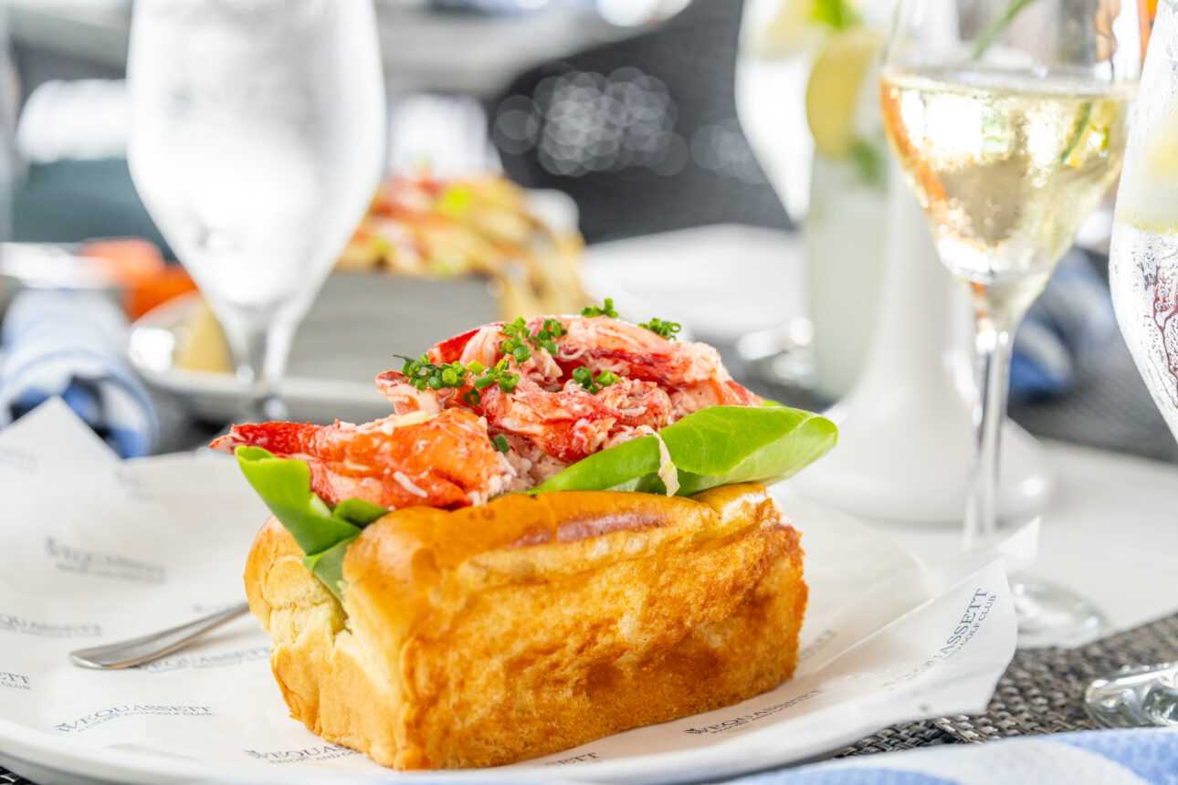 A lobster roll on a piece of parchment paper is served next to two glasses of white wine and a glass of water on a dining table.
