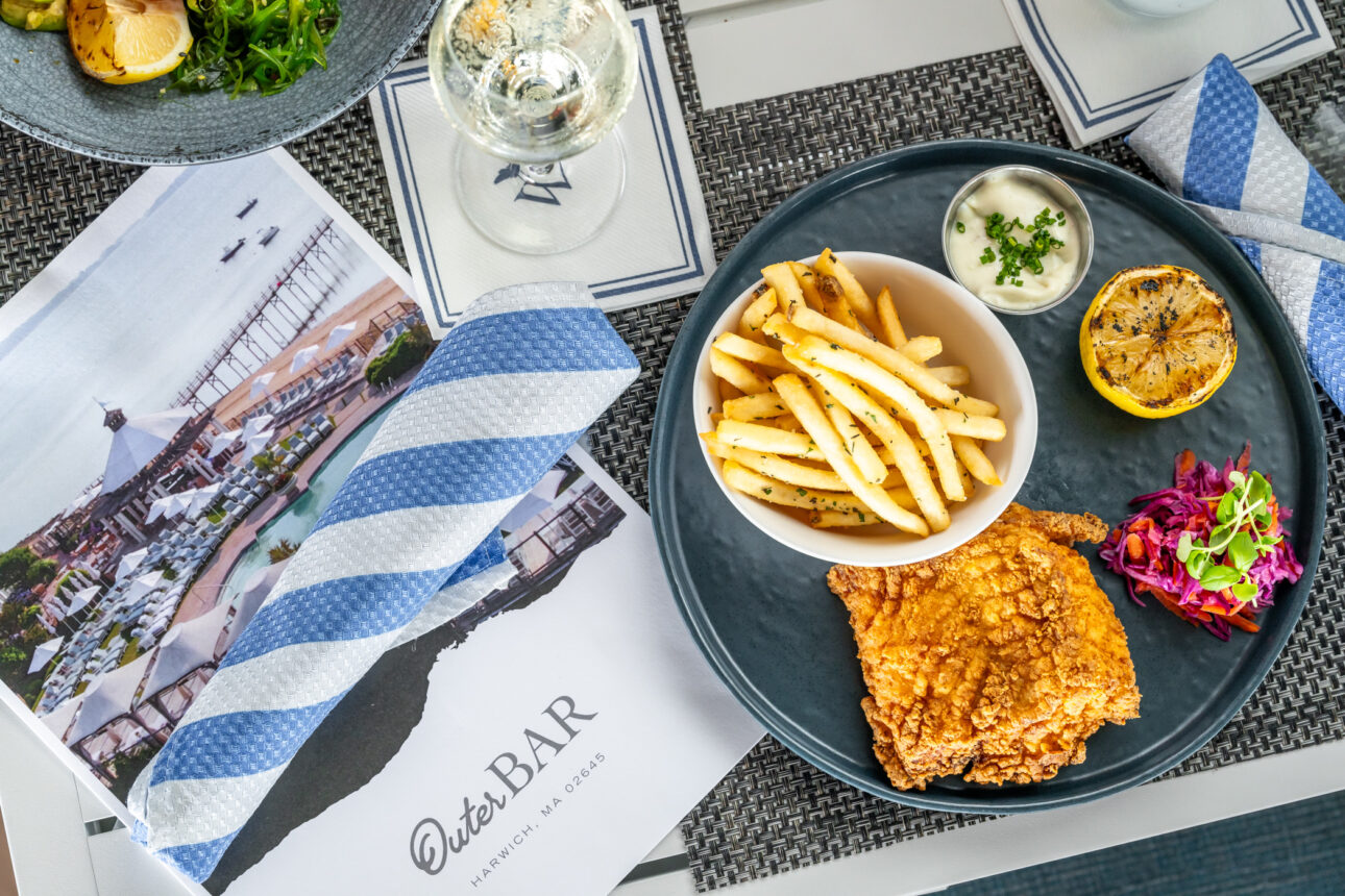 A plate of fried fish with french fries, coleslaw, a grilled lemon, and tartar sauce is served next to a glass of white wine.