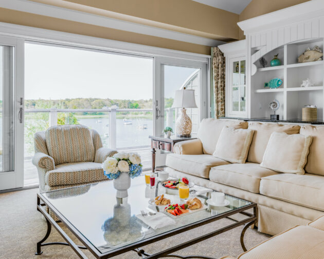A living room with a beige sofa, patterned armchair, glass coffee table with snacks and drinks, and a large sliding glass door offering a view of a lake.