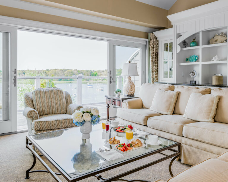 A living room with a beige sofa, patterned armchair, glass coffee table with snacks and drinks, and a large sliding glass door offering a view of a lake.