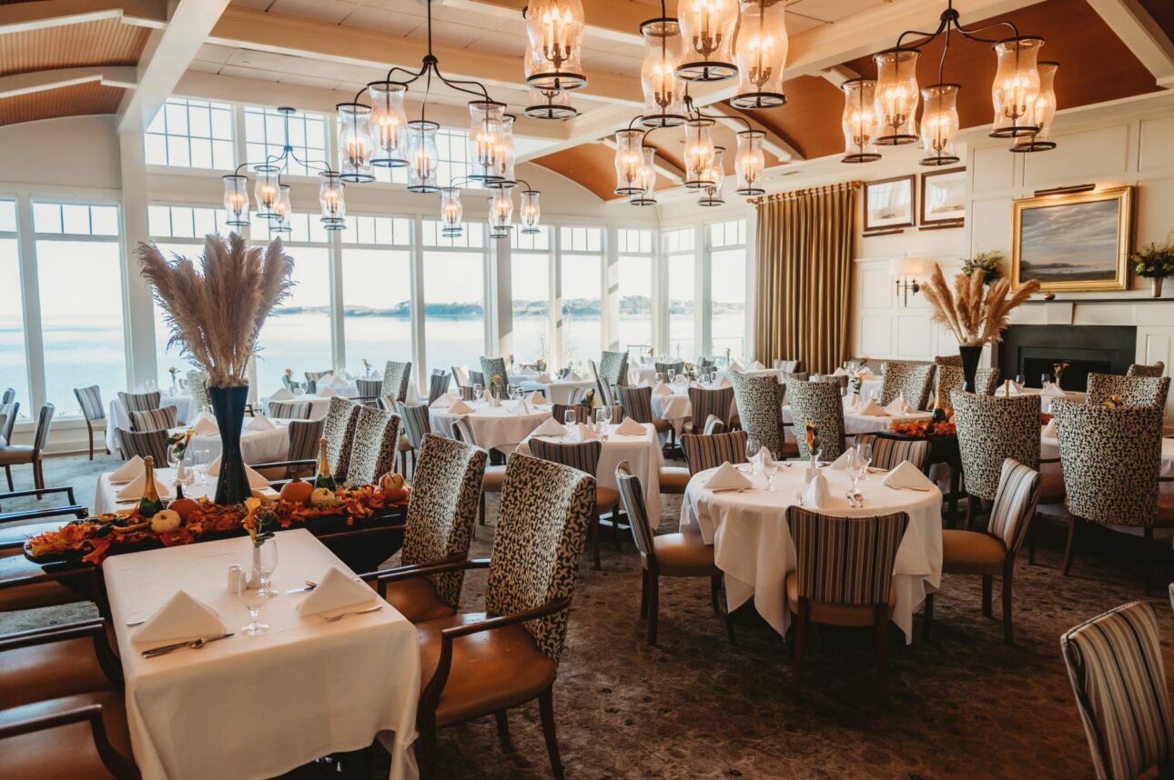 Elegant dining room with neatly set round tables, wooden chairs, decorative lamps, and large windows offering a scenic view of water and hills in the background.
