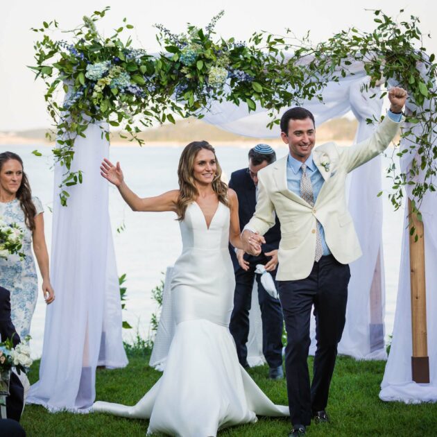 A bride and groom celebrate while walking down the aisle after their outdoor wedding ceremony by the water, surrounded by bridesmaids, groomsmen, and guests.