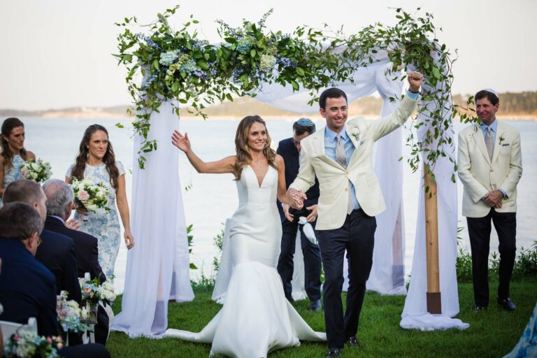 A bride and groom celebrate while walking down the aisle after their outdoor wedding ceremony by the water, surrounded by bridesmaids, groomsmen, and guests.