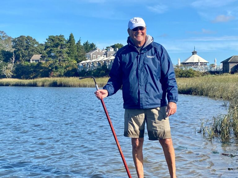 A man in a blue jacket and beige shorts stands in shallow water near a grassy shoreline, holding a red walking stick.