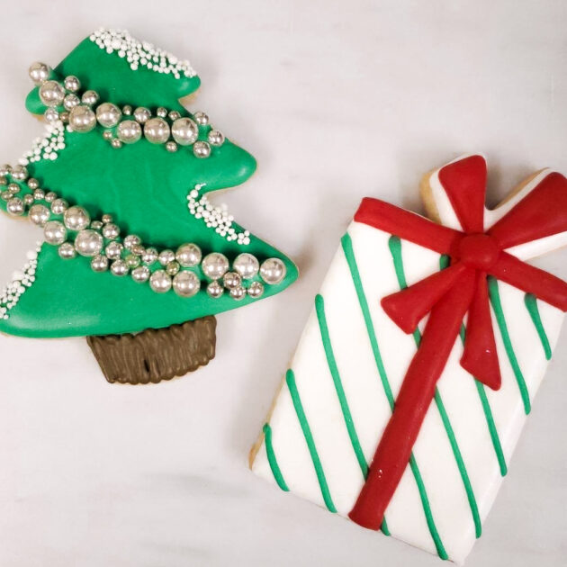 Two decorated Christmas cookies: a green Christmas tree with silver beads and a white gift box with green stripes and a red bow on a light-colored surface.
