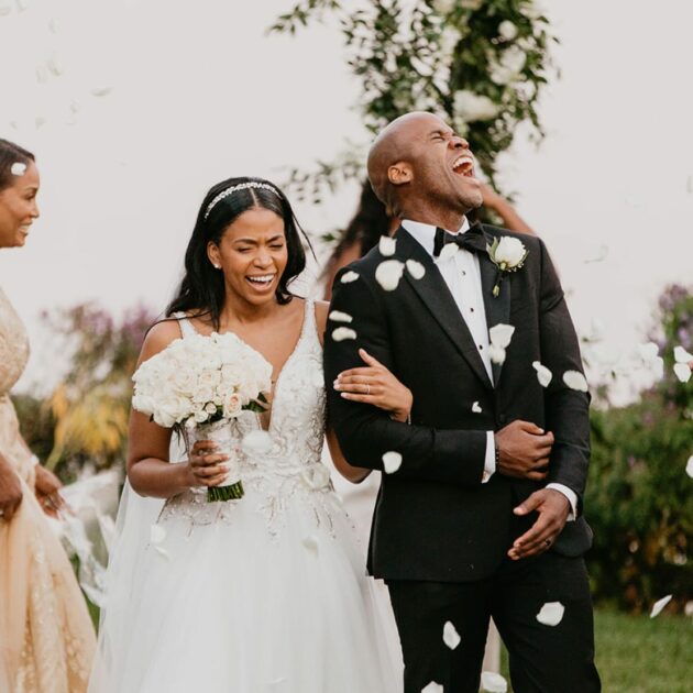 A couple in wedding attire is celebrating outdoors with confetti falling around them. The bride holds a bouquet, while the groom is laughing and holding her arm.