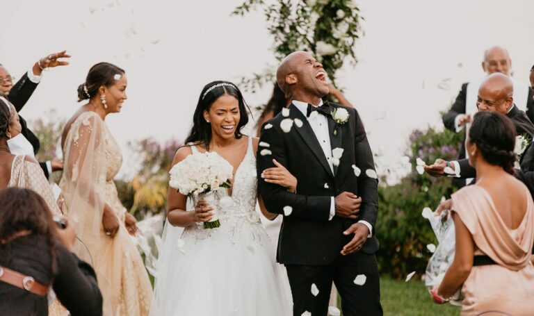 A couple in wedding attire is celebrating outdoors with confetti falling around them. The bride holds a bouquet, while the groom is laughing and holding her arm.