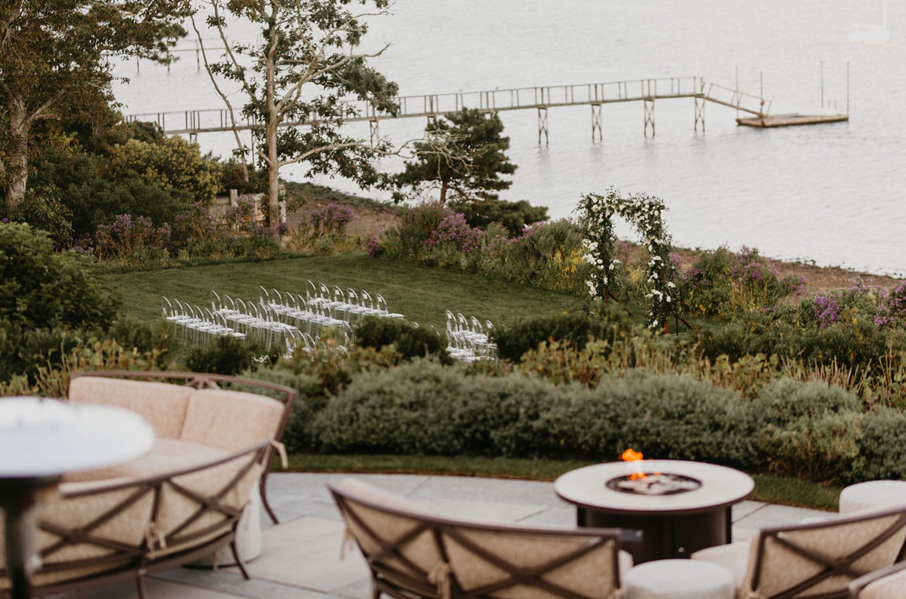 A waterside outdoor wedding setup with white chairs arranged on a lawn, surrounded by greenery. A dock extends into the water in the background.