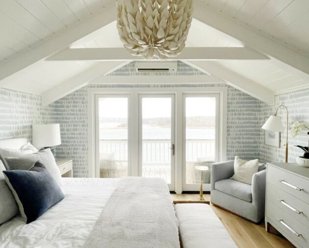 A cozy, light-filled bedroom features a bed, armchair, dresser, and a large chandelier under a vaulted ceiling, with glass doors opening to a balcony overlooking a water view.