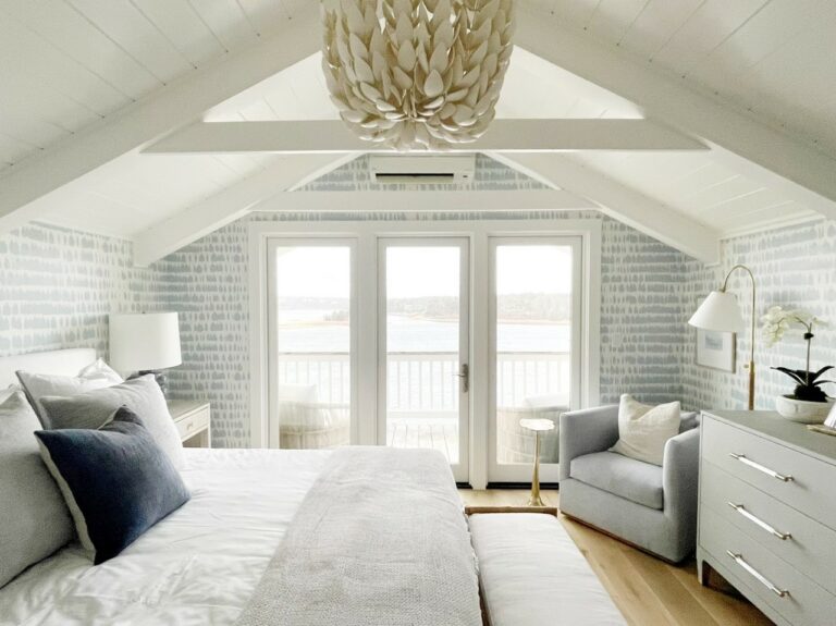A cozy, light-filled bedroom features a bed, armchair, dresser, and a large chandelier under a vaulted ceiling, with glass doors opening to a balcony overlooking a water view.