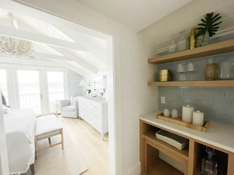 A bright bedroom with white decor is visible from a hallway, featuring open shelves holding decorative items. The bedroom has a large window, white dresser, and a sitting area with a chair.