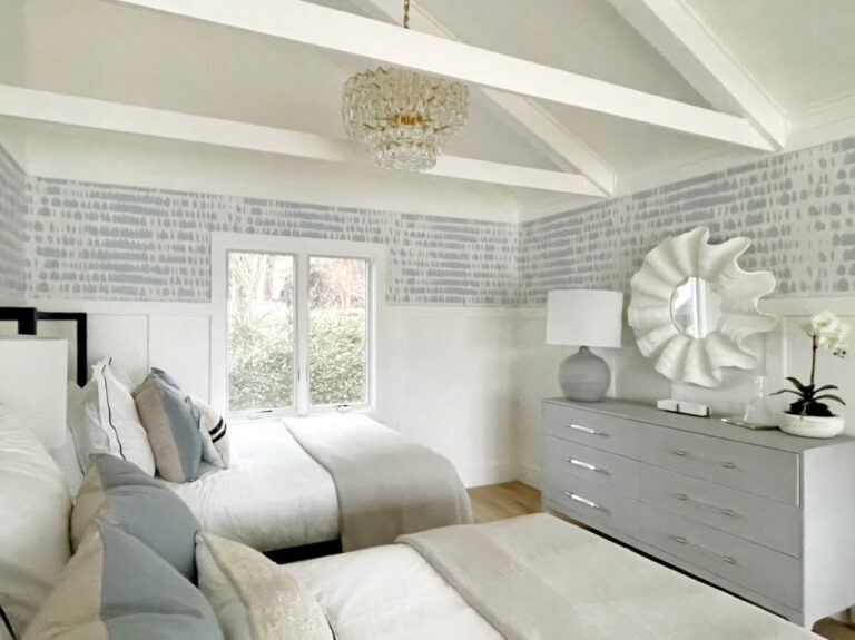 A bedroom with two twin beds, a chandelier, a dresser with a mirror above, a table lamp, and a small orchid plant. The walls have light blue patterned wallpaper, and there are exposed ceiling beams.