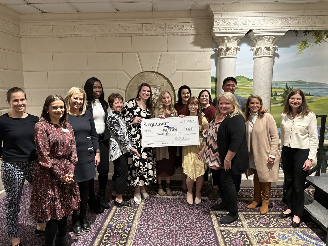 A group of people stands indoors holding a large check from Wequassett. The setting includes a painted backdrop and columns, with a patterned carpet on the floor.