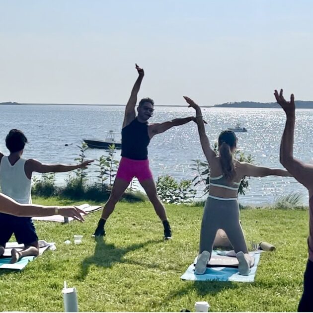 Isaac Boots teaching a workout class near a body of water, with one person standing and others kneeling on mats while stretching their arms.