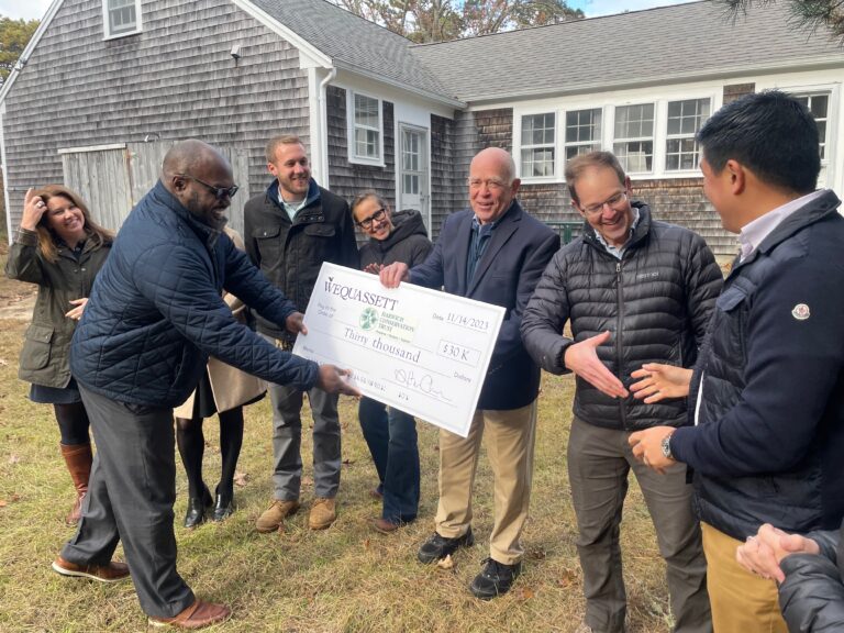 A group of people stands outdoors, some holding a large ceremonial check for $30,000. They are smiling and appear to be celebrating.