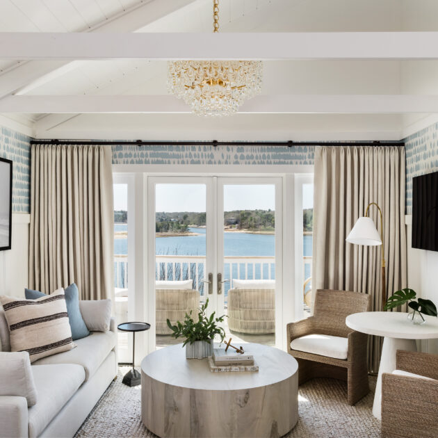 A brightly lit room in the Hydrangea Cottage with white walls, a light-colored sofa, chairs, a round coffee table, a large wall-mounted TV, and a window with curtains overlooking a body of water.