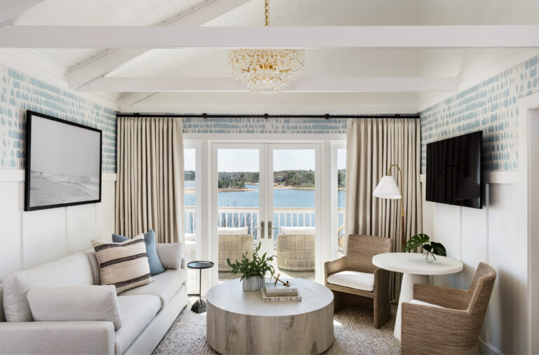 A brightly lit room in the Hydrangea Cottage with white walls, a light-colored sofa, chairs, a round coffee table, a large wall-mounted TV, and a window with curtains overlooking a body of water.