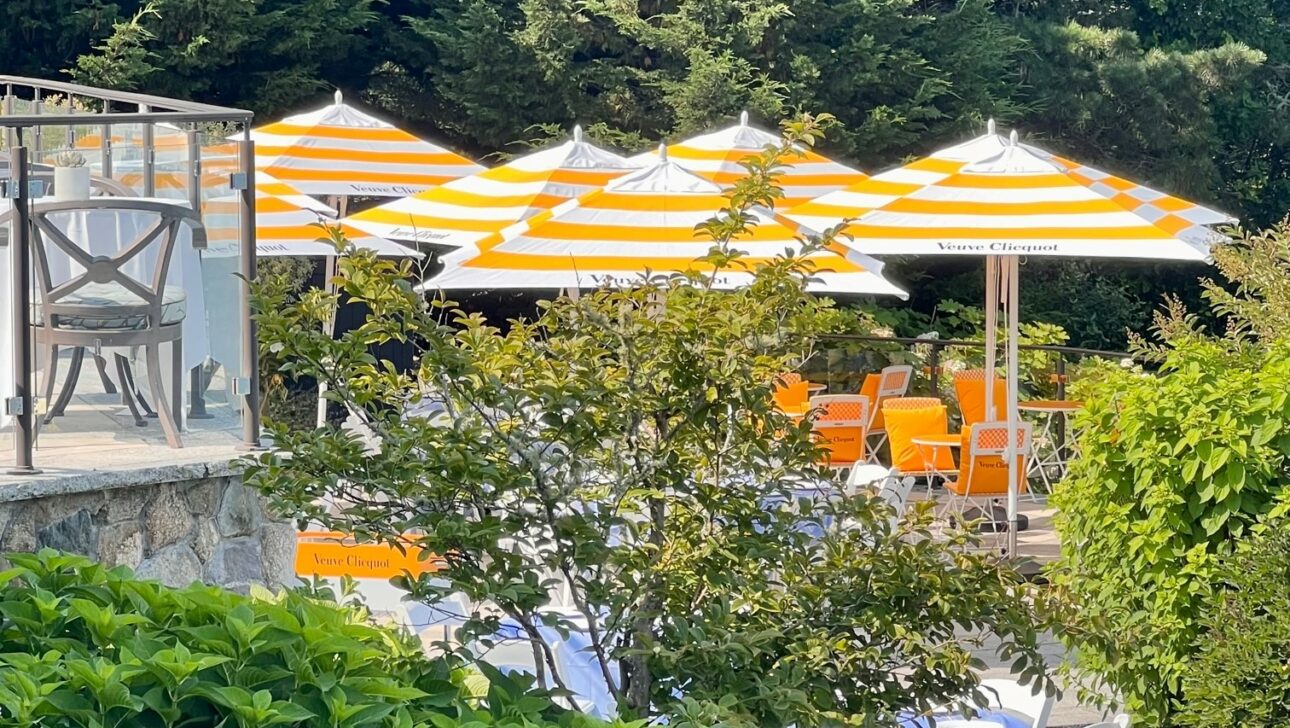 Outdoor seating area with several tables and chairs, shaded by large white and yellow striped umbrellas.