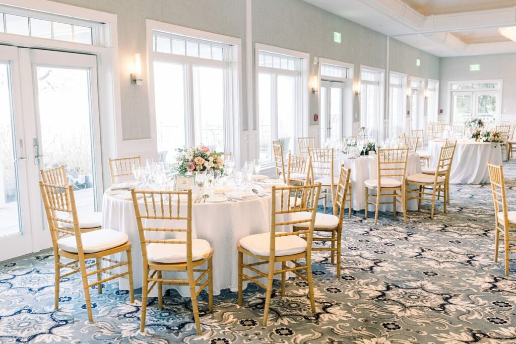 A bright, elegant event space with several round tables covered in white tablecloths, set with glassware and floral centerpieces. Wooden chairs surround the tables, and large windows provide natural light.