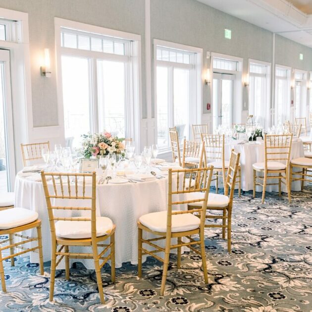 A bright, elegant event space with several round tables covered in white tablecloths, set with glassware and floral centerpieces. Wooden chairs surround the tables, and large windows provide natural light.