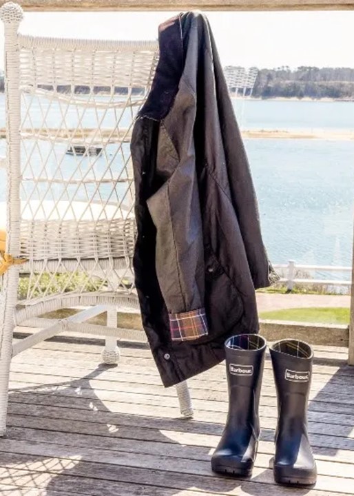 A black Barbour jacket hangs on a white wicker chair on a wooden deck next to a pair of black rubber boots with water and trees visible in the background.