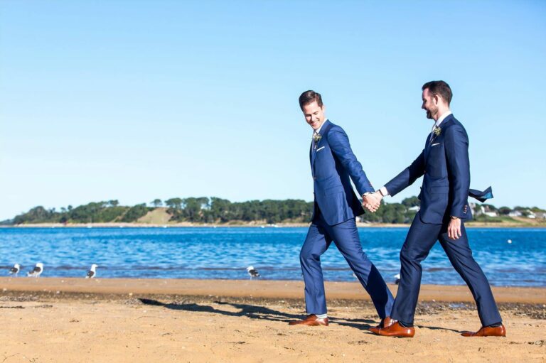 Two men holding hands and walking on the beach.