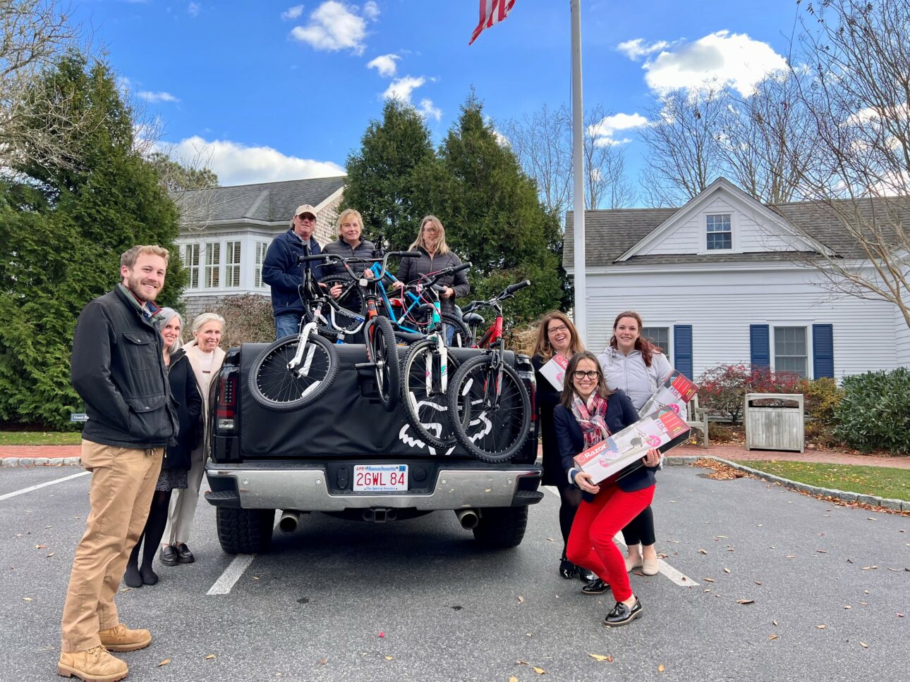 A group of people stand around a truck loaded with bicycles and holiday items in a parking lot, in front of a building, with an American flag in the background.