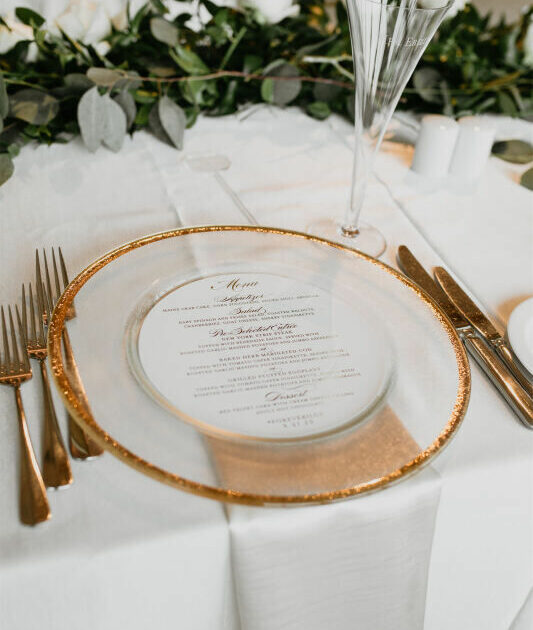 A place setting with a clear charger edged in gold, containing a printed menu. Silverware and a champagne flute are positioned alongside, with greenery in the background.