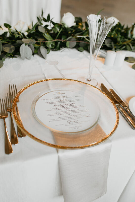 A place setting with a clear charger edged in gold, containing a printed menu. Silverware and a champagne flute are positioned alongside, with greenery in the background.