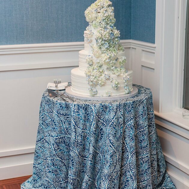 A three-tiered white wedding cake adorned with cascading flowers on a table with a blue patterned tablecloth, situated in a corner of a room with blue walls and white trim.