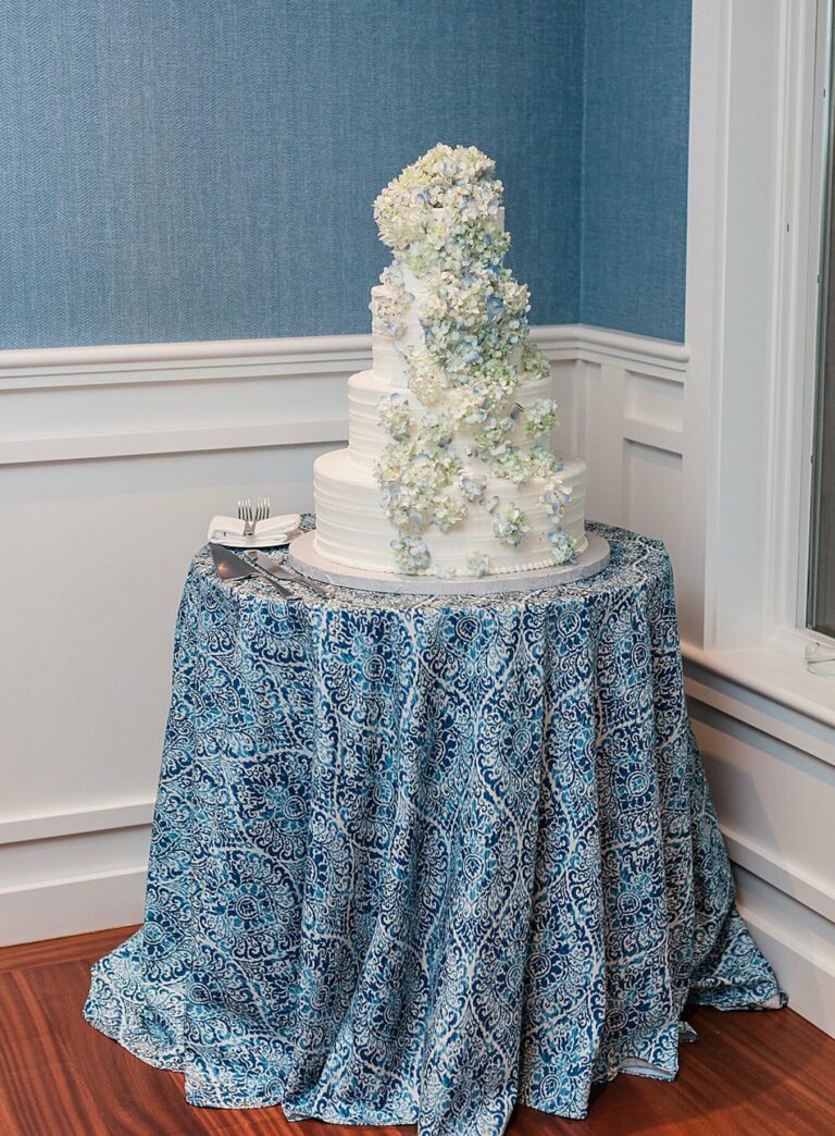 A three-tiered white wedding cake adorned with cascading flowers on a table with a blue patterned tablecloth, situated in a corner of a room with blue walls and white trim.