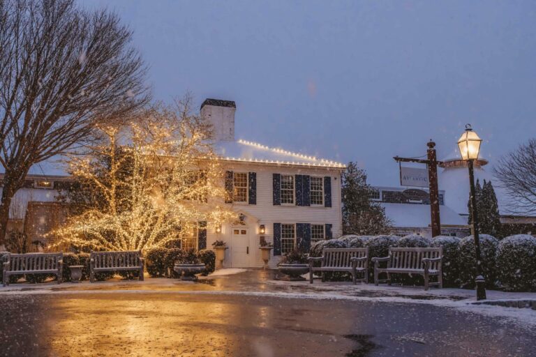 A large white house adorned with festive string lights around the windows and a tree, set against a snowy evening backdrop with benches and a lit street lamp.