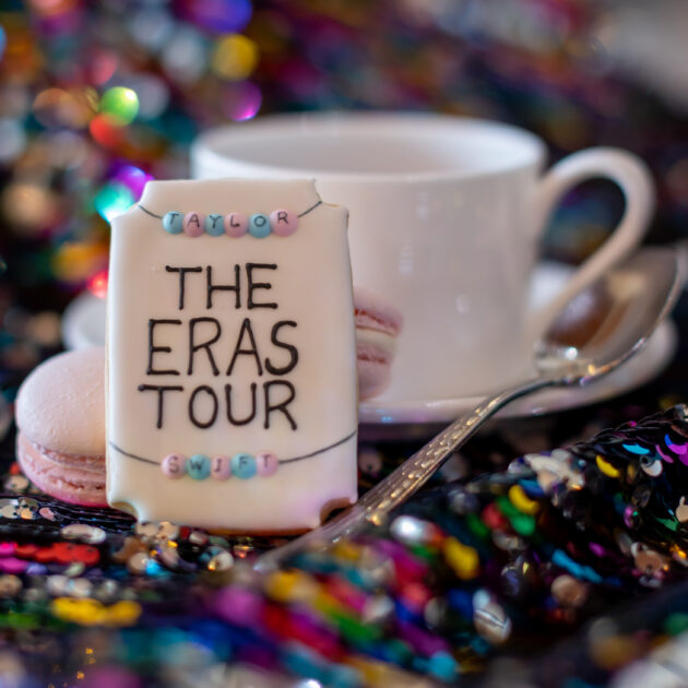 A decorated cookie with the text "The Eras Tour" on it, placed next to a teacup, a spoon, and macarons against a colorful, sequin-studded background.